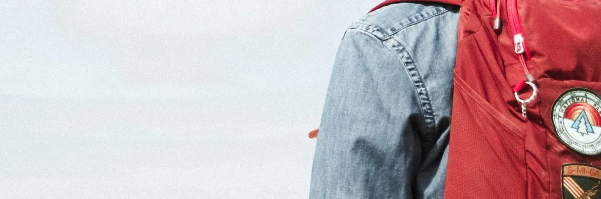 A close-up of a traveler wearing a denim jacket and a red backpack adorned with outdoor-themed patches.